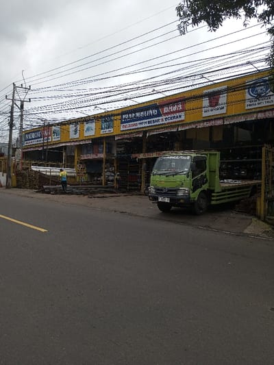 The image captures an outdoor scene featuring a building materials supply store situated alongside a paved road under an overcast sky.
In the foreground, a dark gray asphalt road runs horizontally across the lower third of the image, marked with a faded yellow line on the left side, indicating a lane divider. The road appears dry.
Dominating the mid-ground is a commercial building, likely a hardware or building supply store, characterized by its bright yellow facade. The building has a long, open-front design, allowing views into the interior where various construction materials are stocked. Overhead, numerous black electrical wires crisscross the sky, running from a utility pole on the far left of the frame towards the right.
Several large signs are displayed prominently on the yellow facade. From left to right, partially visible signs include "PANEMBONG." A central, larger sign clearly reads "PANEMBUNG DAJA." Below this, smaller text states "MELAYANI PARTAI BESAR & ECERAN," which translates to "SERVING LARGE PARTIES & RETAIL." Further to the right, another sign displays "BESI BETON SNI" (referring to SNI standard concrete iron or rebar), and another sign below it reads "BAJA RINGAN" (light steel) with "PRIMA INTI TRUS" underneath.
In front of the store, on the paved lot, various building materials are visible. On the left side, there are stacks of pipes and other cylindrical materials, some dark, some lighter. A person wearing a bright yellow or green high-visibility vest and dark pants is standing among these materials, appearing to be working or arranging them.
A green flatbed truck with a white cab is parked to the right of the person, partially under the store's overhang. The truck's license plate, "F 7853 DA," is visible on its front bumper. Text on the truck's windshield or sun visor appears to read "SMILLER." The truck's flatbed is empty.
The sky above is uniformly gray and cloudy, suggesting an overcast day. Trees and foliage are visible in the upper right corner, partially obscured by the electrical wires. The overall impression is that of a bustling commercial area, likely in a developing urban or semi-urban setting.