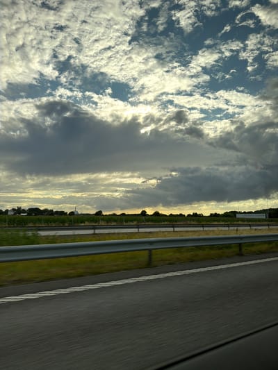 This is a photograph taken from a vehicle traveling on a road, likely a highway, in Åskloster, Sweden. The image captures a wide view of the sky and the surrounding landscape.
The sky is filled with a dramatic cloud formation, with a mix of dark, heavy clouds and brighter, illuminated patches suggesting sunlight breaking through. The clouds appear textured and dynamic, with some areas showing a silvery sheen. The upper portion of the sky reveals patches of blue.
Below the sky, a horizontal band of light stretches across the horizon, likely representing the setting or rising sun, casting a warm glow. In the middle ground, a line of trees and some buildings are visible, suggesting a rural or semi-rural area. Among these structures, a tall industrial chimney with some white markings is noticeable. A vineyard or a field with rows of plants is also visible in front of the treeline.
In the foreground, a metal guardrail runs parallel to the road, separating the lanes of traffic. The road surface, visible in the lower part of the image, is dark asphalt with white lane markings. The motion blur on the road and the guardrail indicates that the photograph was taken while the vehicle was in motion. The overall impression is of a scenic drive under a moody sky.