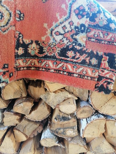 A close-up, high-angle shot shows a section of an old, worn rug with a faded orange background and a repeating pattern of black, cream, and blue floral motifs. The rug is draped over a pile of neatly stacked firewood. The logs are of various sizes, predominantly birch, with visible bark and wood grain. The scene is outdoors, possibly in a rural setting or backyard. The lighting suggests daytime, with a warm, natural light illuminating the textures of the rug and wood. The specific location of Lobkovo, Russia is not evident in the visual details of the image.