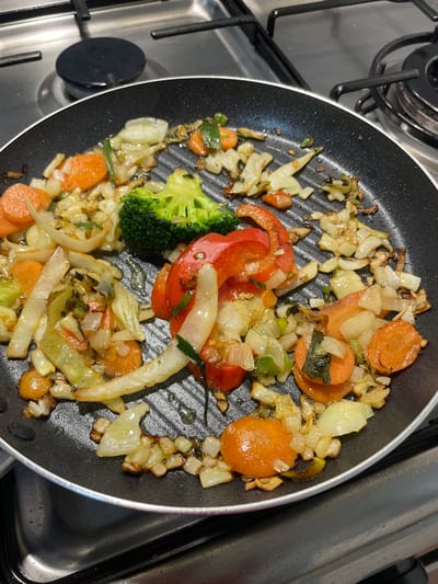 The image displays a close-up, high-angle view of various sautéed vegetables in a black non-stick grill pan, positioned on a stainless steel gas hob. The vegetables, distributed unevenly across the pan's ridged surface, include sliced carrots, chopped white and red onions, a distinct floret of broccoli, and sliced red bell pepper. Some vegetables exhibit browning and caramelization, indicating partial cooking, and residual oil droplets are visible in the pan's grooves. In the background, sections of two black cast-iron burner grates and a black burner cap are discernible on the stainless steel surface of the stovetop. The scene is located in a kitchen in Covilhã, Portugal.