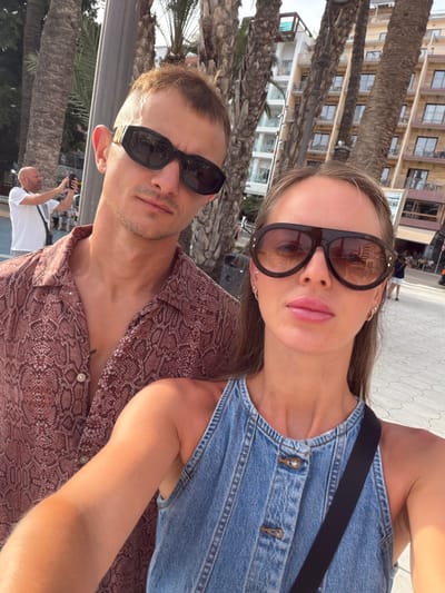 The image is a selfie taken by two people, a woman and a man, in an outdoor setting with palm trees and buildings visible in the background. The woman is in the foreground, wearing sunglasses and a denim garment. The man is beside her, also wearing sunglasses and a patterned shirt. In the background, two other people are visible, one of whom appears to be holding a camera. The scene suggests a sunny day, possibly in a tourist area, given the presence of palm trees, buildings, and other people. There is no visible text in the image. The overall mood appears casual and relaxed, consistent with a vacation or leisure activity. The location context suggests Benidorm, Spain.