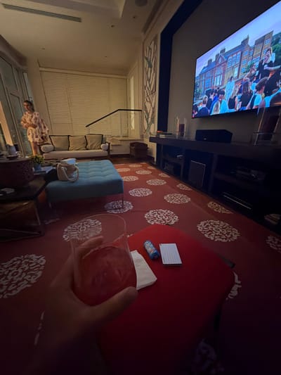 The scene depicts an interior living space in La Romana, Dominican Republic, viewed from a low perspective. The room is dimly lit, suggesting evening. In the immediate foreground, a human hand holds a clear glass containing a reddish liquid with ice. This hand rests above a low, square, red ottoman or table. On this red surface, a small white napkin, a blue and white cylindrical item (possibly a can), and a stack of small white notepaper are visible.
The floor is covered by a red carpet featuring a repeating white circular or floral pattern. Centered in the mid-ground is a rectangular ottoman upholstered in light blue or teal fabric with metal legs. To the left, a dark-colored rectangular coffee table is partially visible.
In the background on the left, a female figure stands near a large window or glass door fitted with horizontal blinds. She is wearing a light-colored, patterned dress or robe. Behind her, a neutral-toned sofa with throw pillows is placed against the wall beneath the blinds. A black, abstract, minimalist wall decoration is mounted to the right of the blinds.
Along the right wall, a long, dark-colored media console holds a large flat-screen television, a soundbar positioned centrally, and other electronic devices within its shelves. The television screen displays a scene featuring numerous people, primarily young adults, gathered outdoors in front of brick buildings, consistent with a movie or television show. The walls are light-colored, and a decorative panel with a muted, abstract branch-like pattern is visible between the window area and the television.
