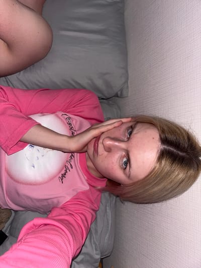 An eye-level indoor close-up selfie captures a young woman looking directly at the camera with a displeased expression. She has blonde hair with prominent dark roots, parted down the middle, and striking blue eyes. Her fair skin displays some redness, freckles, and minor blemishes. Her left hand supports her chin and cheek, emphasizing her furrowed brows and downturned mouth, conveying an emotion of annoyance, boredom, or slight frustration.
She is wearing a long-sleeved pink shirt with ribbed sleeves. The front panel of the shirt, which is a lighter pink, features visible black cursive text that reads "Crystal School Board" alongside a faint graphic, possibly a cat's face or other animal silhouette within a circular outline.
To the woman's left, a gray pillow is visible, and in the bottom left corner, a bare leg or knee is partially seen, suggesting she is in a relaxed, possibly seated or lying position on a bed or sofa. The background consists of a light-colored, textured wall or ceiling with a diagonal linear pattern, likely wallpaper. The lighting is soft and even, typical of an indoor setting. The scene is located in Ligolambi, Russia.
