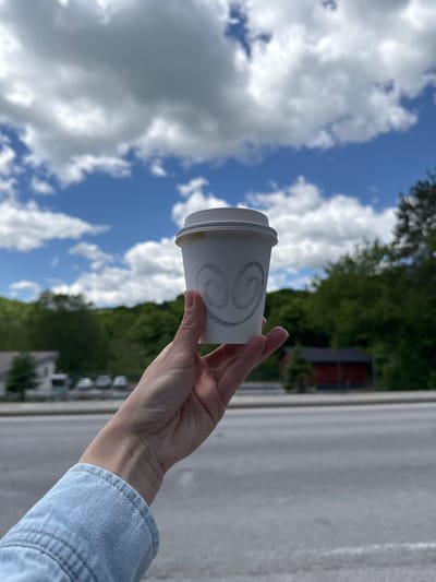 A person's hand holds a white paper coffee cup with a swirling design against a backdrop of a partly cloudy blue sky and green trees. A gray asphalt surface and blurry buildings suggest an urban or suburban environment. While the general scene is provided, a specific location in Chelsea, Canada, is not discernible from the visual information.