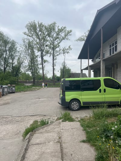 The image shows a bright green van parked on a concrete street in what appears to be an urban or suburban area of Cherliany, Ukraine. The van is parked next to a curb with grassy areas on the other side, interspersed with dandelion-like plants. A building with multiple stories and unfinished construction is visible behind the van. Trees are in the background, and two individuals are walking in the distance. Several trash bins are visible along the street. The sky is overcast.