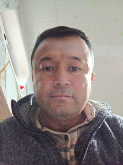The image is a headshot-style selfie taken indoors, depicting an adult male, identified as being in the city of Suti, Uzbekistan. The man, appearing to be middle-aged, has short, dark hair, a light to medium skin tone, and stubble on his face. He is looking directly at the camera with a neutral expression. He is wearing a light-colored plaid shirt with dark buttons, under a dark grey or charcoal-colored knitted hooded cardigan or jacket. The collar of the plaid shirt and the inner lining of the cardigan collar are visible. The background consists of an interior wall and ceiling. To the left, a light-colored vertical panel or doorframe is partially visible, alongside what appears to be a patterned or textured wall surface in light tones. The ceiling is white with scattered, small, light-colored (possibly green or multi-colored) star-like patterns or specks. No other individuals, distinct objects, or explicit actions beyond the self-portrait are observable.