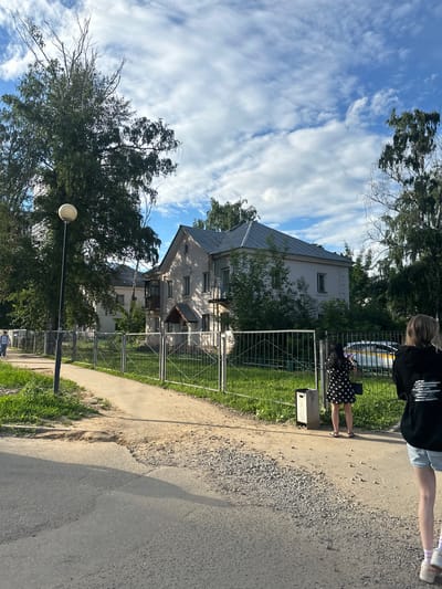 A vertical shot captures a suburban scene in Kotelniki, Russia, on a bright, cloudy day. Two individuals are partially visible in the foreground. On the left, a person with blonde hair wears a black t-shirt and blue shorts. To the right, a person in a black polka dot dress stands near a fence. The background features a multi-story building with a grey roof, surrounded by lush green trees. A metal fence encloses a grassy area. The ground is a mix of asphalt and gravel. A lamppost is visible on the left side of the image. The sky is a vivid blue, with streaks of white clouds. Sunlight casts long shadows, indicating it is likely late afternoon. No discernible text is visible.