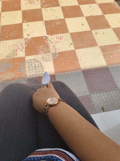 A person sits with dark gray pants and a rose-gold wristwatch on their arm. The ground is a checkered pattern of red-brown and white squares, littered with small debris. A white sock is partially visible in the upper part of the image. The shot is taken from a slightly elevated perspective. A hint of a patterned garment peeks into the bottom of the frame.