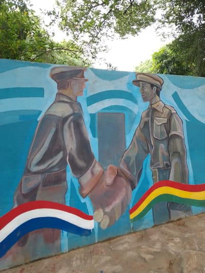 The image depicts a mural in Villa Hayes, Paraguay. Two men in military-style uniforms are shaking hands. The background is a blue sky with white stripes. A Dutch flag is painted on the left, while the Bolivian flag is on the right. The mural is located on a concrete wall outdoors. Trees and sky are visible in the background.