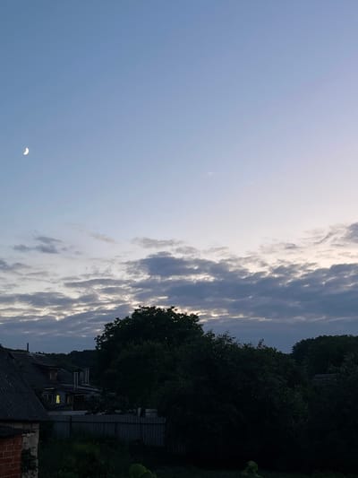 This is a vertical, medium shot taken from an elevated viewpoint, looking out over a rural or suburban landscape at dusk. The sky occupies the upper two-thirds of the frame and is a gradient of pale blue to light grey, with a sliver of the crescent moon visible in the upper left quadrant. A band of textured, grey clouds stretches across the middle ground of the sky.
The lower third of the image depicts a dense canopy of dark green trees, silhouetted against the brighter sky. In the left foreground, the dark, weathered roof of a building, possibly a house, is visible, constructed with slate tiles and some exposed brickwork. To the right of this building, another structure with a sloping, dark roof and visible chimneys is partially illuminated by a single, warm yellow light emanating from a window. A corrugated metal fence runs horizontally across the middle ground, separating the foreground from the background. The overall impression is one of a tranquil evening scene in the countryside near Krasnokutsk, Ukraine, with the transition from day to night.