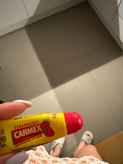 The image shows a close-up of a person's hand holding a tube of Carmex strawberry flavored moisturizing lip balm. The lip balm tube is yellow with red lettering and a graphic of strawberries. The background appears to be a tiled floor with shadows, suggesting an indoor setting. There are also feet visible in the lower right of the image wearing striped slippers. The presence of what appears to be a shower curtain rod and tiled walls in the upper left corner suggests the location might be a bathroom. There is no visible indication that this scene is located in the city of Murcia, Spain.