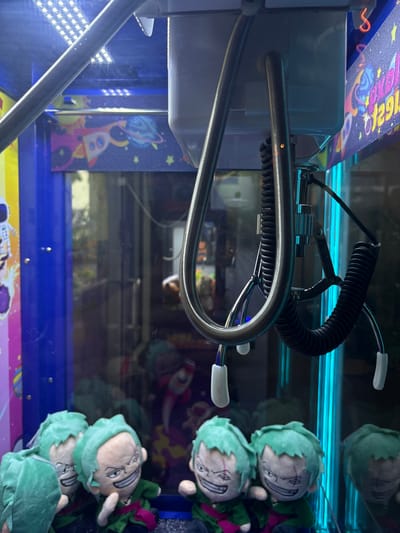 The image shows the interior of a claw machine, a type of arcade game. The machine contains several plush toys, all appearing to be characters with green hair and distinctive facial markings, likely from an anime series. The claw mechanism, made of shiny metal and coiled black tubing, is visible at the top of the frame, poised above the prizes. The background of the machine features colorful, space-themed decorations, including rockets and planets. The lighting is predominantly blue, with some white LEDs illuminating the top section. There are no people visible in the image, and no specific location in Orlando, United States of America can be identified from the visual cues.