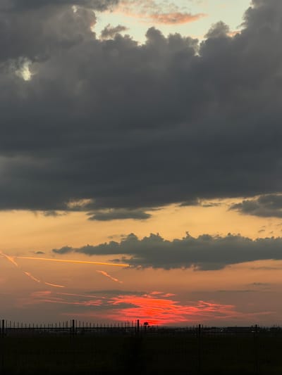 The image depicts a sunset over a rural landscape, possibly in or near the village of Timofeyevka, Russia. The sky is filled with dramatic clouds, transitioning from dark gray at the top to vibrant orange and red hues near the horizon. Multiple contrails are visible, streaking across the sky, some illuminated by the setting sun. In the foreground, a dark silhouette of a metal fence runs horizontally across the bottom of the frame, with a field of dark vegetation behind it. Beyond the field, a faint outline of industrial or agricultural structures can be seen against the brightly colored sky. The overall scene conveys a sense of atmospheric drama and the fading light of day. There are no people or discernible animals present.