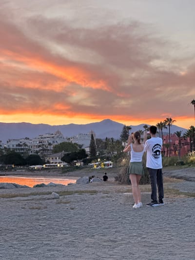 Two people are standing on a beach in El Velerín, Spain, taking pictures of a sunset over the Mediterranean Sea. The sky is filled with vibrant orange and pink clouds. In the background, there is a town with white buildings and palm trees. The sea reflects the colors of the sunset. The beach is sandy and there are rocks scattered on the shore. The people appear to be a couple, both looking out at the view and holding up their phones to capture the moment. The man is wearing a white t-shirt with a graphic print and dark pants, while the woman is wearing a white tank top and a green skirt. The overall scene is peaceful and romantic, capturing the beauty of a Spanish coastal sunset.