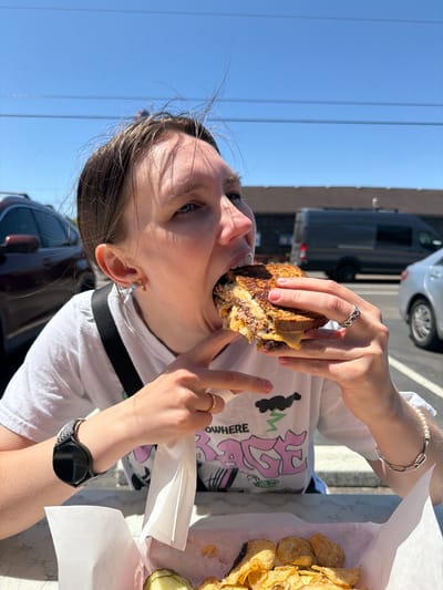 A young woman with brown hair pulled back eats a sandwich outdoors, against a backdrop of a bright blue sky with power lines. She is wearing a white t-shirt with a graphic design, a black watch on her left wrist, and jewelry on her fingers and right wrist. Her mouth is open as she bites into the sandwich. In front of her is a basket of potato chips and some pickle slices. In the background, a dark gray van and a silver car are parked. The image appears to be taken on a sunny day in Cannon Beach, Oregon, United States.