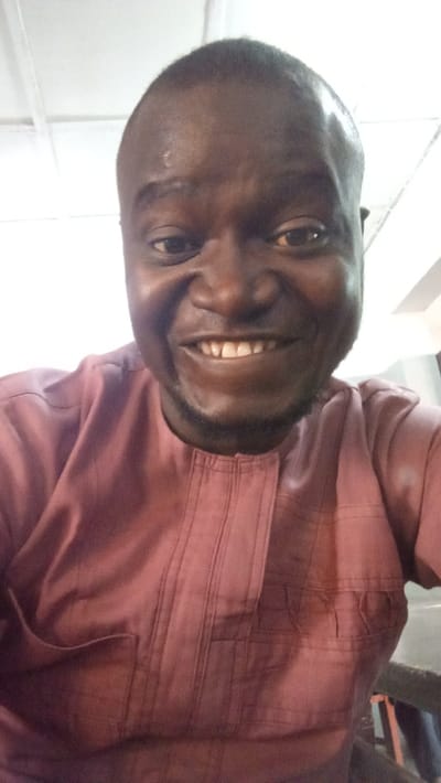 The scene is situated indoors in Dakata, Nigeria. A dark-skinned adult male is depicted in a close-up, frontal portrait. He has a bald head and faint stubble along his jawline. His face displays a broad smile, revealing upper and lower teeth. His eyes are dark and open, directed towards the viewer. He is wearing a reddish-brown traditional garment, possibly a tunic, featuring vertical stitch lines or pleats on the chest area and a rectangular pocket on the left side of the torso. The background consists primarily of a bright, white ceiling with visible grid lines and panels, indicative of an interior space. Faint, darker elements are present behind the man's shoulders, suggesting indistinct architectural features or furniture in the periphery.