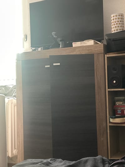 A room in Gelsenkirchen, Germany, features a large television screen mounted above a cabinet. The cabinet has a dark, textured front with two handles. To the right of the cabinet is a shelving unit containing a speaker and some boxes. A radiator is visible to the left of the cabinet, partially obscured by a gray blanket. The room is lit by natural light coming from a window.