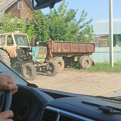 An old, rusty blue and orange tractor is parked next to a rusty brown trailer on a dirt road. The tractor has its hood open, revealing the engine. Trees with green foliage and some yellowing leaves are behind the tractor and trailer. A wooden building with a peaked roof and a small window is visible to the left. To the right, a light blue metal fence runs along the property line, with a concrete utility pole in front of it. A yellow line is painted on the dirt road in the foreground. The image is taken from inside a vehicle, with a steering wheel and dashboard visible. The sky is clear and blue.