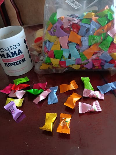 A close-up, top-down shot captures a collection of colorful, individually wrapped candies scattered across a dark brown, glossy surface, likely a table. The candies are in various fruit-themed wrappers: purple with grapes, pink with peaches, yellow with what appears to be citrus, orange with possibly oranges or apricots, and blue with an unspecified fruit. Some of the wrappers have embossed images and Chinese characters.

To the left, a white ceramic mug is visible, partially obscured by a transparent plastic bag. The mug features Cyrillic text, including "крутая МАМА" (cool MOM) and "ПОТРЯСАЮЩЕЙ ДОЧЕРИ" (for an amazing DAUGHTER). Below this, smaller text reads "Да, это она подарила мне эту кружку" (Yes, she gave me this mug).

Behind the mug and to the right, a large, clear plastic bag is filled with an abundance of similar candies, creating a vibrant mosaic of colors. The bag is open at the top. The overall impression is a still life of sweets, possibly a gift or a treat, placed on a table. The scene is indoors, with soft lighting that highlights the glossy surfaces of the candies and the table. The setting does not provide specific visual cues to determine the precise location within Brodokalmak, Russia, beyond suggesting an indoor domestic environment.