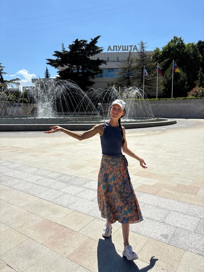 In Alushta, Ukraine, a woman stands in a paved plaza with a fountain and a building in the background. She is wearing a blue sleeveless shirt, a colorful wrap skirt, a white baseball cap, and white clogs. Her left arm is extended to the side with her palm up, as if presenting something. The fountain sprays water upwards behind her, and the building behind the fountain has the word "АЛУШТА" in large letters on its facade. Three flags, including the Russian flag, fly in front of the building. The ground is paved with large, light-colored square tiles.