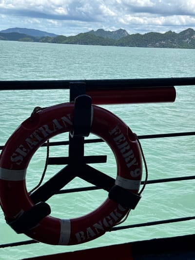 This image depicts a red life preserver with white lettering, attached to a black metal railing on what appears to be a ferry. The words "SEATRANS FERRY 9 BANGKOK" are visible on the life preserver. In the background, a body of calm, greenish water stretches towards a chain of lush, green islands under a cloudy sky. The scene is likely taken from the deck of a ferry in the waters surrounding Thong Nian, Thailand, a location known for its scenic islands and ferry services.