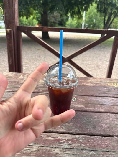 In Gresovsky, Ukraine, a close-up shot captures a hand making the "rock on" gesture next to a clear plastic cup filled with iced coffee and a blue straw. The cup sits on a weathered wooden picnic table, with a blurred outdoor scene of trees and a wooden fence in the background. The image appears to be taken during the daytime.