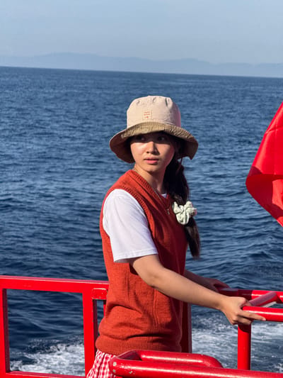 A young East Asian woman with long dark hair tied in a ponytail stands on the deck of a red boat, holding onto a red railing. She is wearing a white t-shirt, an orange vest, a beige bucket hat, and red and white checkered shorts. A light green scrunchie adorns her ponytail. In the background, the deep blue sea stretches to the horizon, with faint mountains visible in the distance. The scene appears to be during daylight, under a clear sky. There are no visible landmarks to suggest the location is specifically in Dong Son, Vietnam.
