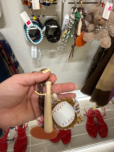 A close-up shot focuses on a person's hand holding a keychain with several charms. The keychain features a miniature baseball glove with a baseball, a wooden baseball bat, a small orange leather tag, and various decorative cords. In the background, other items are displayed on hooks or shelves, including bracelets, a teddy bear keychain with a pearl necklace, and several pairs of red sandals. The overall setting appears to be a retail environment, possibly a clothing or accessories store. There is no information within the image that specifies a location in the city of Leganés, Spain.