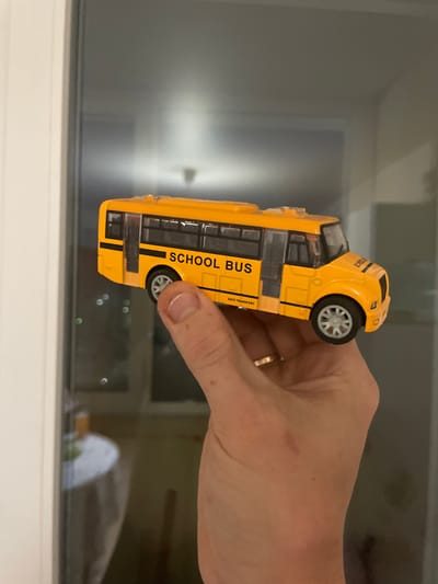 A person's hand holds a miniature yellow school bus model. The bus is detailed with black windows and the words "SCHOOL BUS" printed on its side in black lettering. The background appears to be a window with reflections of lights and a room. The image is taken indoors. There is no information about the city of Laishevo, Russia in this image.
