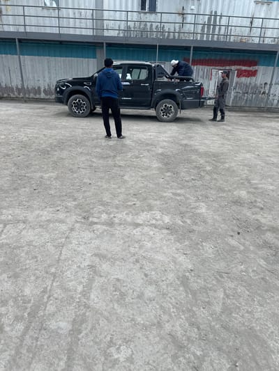 A black pickup truck is parked on a dirt surface in front of a light-colored building with horizontal blue and red stripes. Two men are standing near the truck. One man, wearing a blue hoodie and black pants, stands on the left side of the truck, facing away from the camera. Another man, wearing a dark jacket and white cap, is positioned in the bed of the truck, bending over. A third man, wearing a gray uniform and a dark cap, stands on the right side of the truck, facing the truck. The ground is covered in loose gravel and dirt, suggesting an outdoor industrial or construction area. The building in the background appears to be modular or prefabricated. This scene is located in the city of Takfan, Tajikistan.