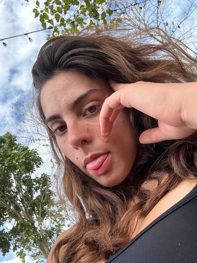 A young woman tilts her head to the side, extending her tongue. Her hand is near her face, partially obscuring her left eye. She has light brown eyes and light brown hair. A mole is visible on her cheek. In the background, branches of trees and a blue sky with clouds are visible. String lights hang among the branches. She is wearing a black tank top.