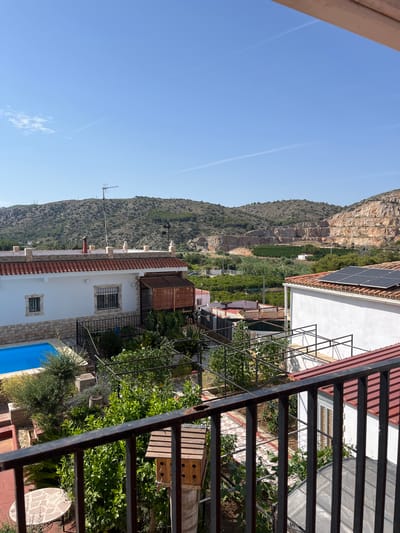 This is a vertical, eye-level, outdoor shot of a residential property in a mountainous region, likely during the daytime on a clear day. The scene is dominated by a steep, rocky mountain face on the left, covered in vegetation in its lower half. Several buildings with terracotta tile roofs are visible nestled into the hillside, surrounded by lush greenery. One building in the foreground is white with stone accents and features a bright blue swimming pool. Solar panels are visible on the roof of another building further up the slope. A metal railing is prominent in the foreground on the right, partially obscuring the view of the property's garden and patio area, which includes a circular mosaic table. A television antenna is mounted on the roof of a building to the left. The overall impression is one of a tranquil, sunny environment in a rural or semi-rural setting, possibly in Spain given the location context. There is no visible text in the image.