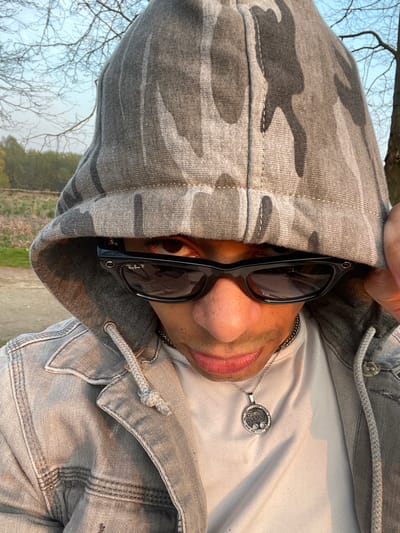 A close-up, eye-level portrait depicts a male subject outdoors in Plaisir, France. The subject is wearing a grey camouflage-patterned hoodie with the hood up, black Wayfarer-style Ray-Ban P sunglasses, a grey denim jacket, and a white t-shirt. A silver-toned chain with a circular pendant, featuring a central emblem and perimeter embellishments, hangs from the subject's neck. The subject's left hand is partially visible near the side of the hood. Warm, soft light illuminates the subject from the right. The background consists of a dirt path, green foliage, and bare trees against a light blue sky, indicative of an open natural area.