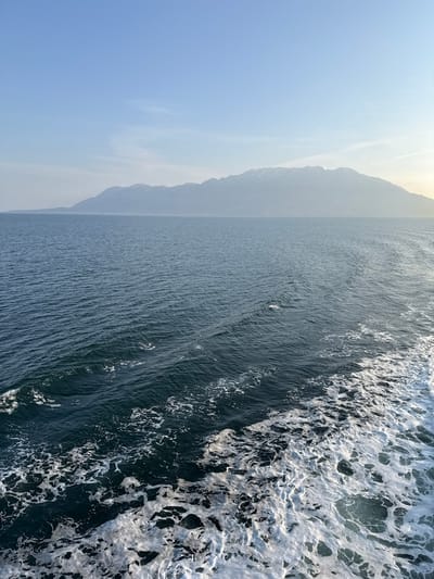 The image depicts a marine scene near Kato Karioutes, Greece, captured from a moving vantage point on the water. The foreground and midground are filled with dark blue-green sea, characterized by numerous ripples and small waves. A significant portion of the lower right displays a thick, white, foamy wake, indicating the recent passage of a vessel. In the distant background, a substantial mountain range extends horizontally across the horizon, appearing somewhat indistinct due to atmospheric haze, with visible patches of snow on its upper slopes. The sky above is clear and light blue, gradually lightening towards the horizon. No individuals or distinct man-made structures are present, save for the implied vessel from which the photograph was taken.