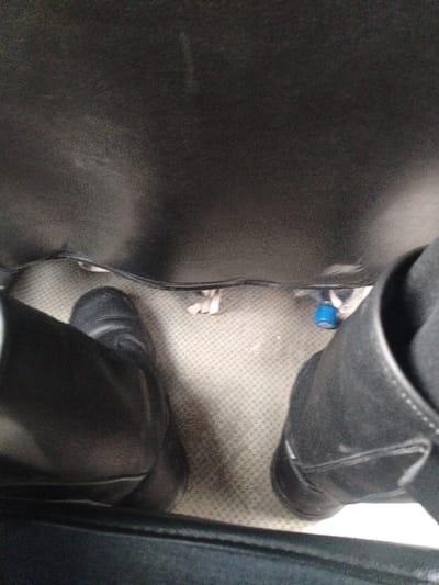 A low-angle view inside what appears to be an airplane cabin. The floor is covered in a beige patterned carpet. Two black leather boots are visible, along with a small blue bottle and some scraps of white material. The seats on either side create a dark, enclosed space. The lighting is dim and casts shadows around the feet and objects on the floor.