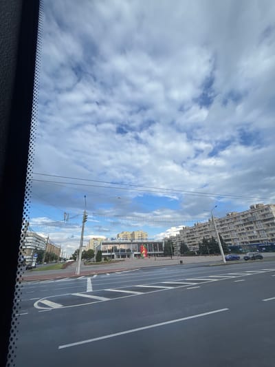 The image shows a wide city street with multiple lanes, marked with white lines. In the foreground, the road surface is dark gray. Beyond the road, there is a sidewalk and a grassy area with trees. Several multi-story buildings are visible, including a large, modern building with glass windows and a sign that appears to be in Cyrillic. A traffic light is present on a pole. Overhead wires are strung across the street. The sky is partially cloudy with patches of blue visible. There are no people clearly identifiable in the image, and no specific actions or interactions are depicted. The scene appears to be in an urban environment. The provided location is within the Senicki Sel'ski Sovet, Belarus.