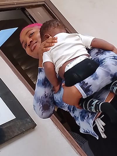 A woman with pink hair is holding a child. The child is wearing a white t-shirt and a diaper with black pants over it. The child is also wearing socks with a pattern. The woman is wearing a blue and white shirt. They are standing in front of a window. The image was taken in Umueshi, Nigeria.