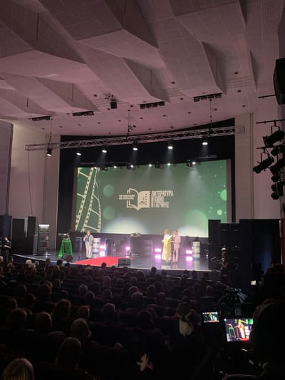 The image depicts an indoor event, likely a ceremony or presentation, taking place in a large hall with a distinct architectural style. The hall features a complex, textured ceiling composed of white, angular panels with a perforated surface, possibly for acoustic purposes. On one side, a large screen displays a green background with subtle bokeh effects and text. The text on the screen appears to be in Russian and reads "ЛИТЕРАТУРА И КИНО В ГАТЧИНЕ" which translates to "Literature and Cinema in Gatchina". Above this text, the number "32" is visible, along with a graphic of an open book and a building.
The foreground of the image shows an audience seated in rows, facing towards the stage area. The audience members are mostly obscured by the darkness and the angle of the shot. The stage itself is illuminated, with several people standing and interacting. Two individuals in the center appear to be presenters or performers, dressed in bright, contrasting outfits – one in yellow and another in a light pink or beige. Other individuals are visible on the stage as well, some seated.
To the right of the stage, there are several large speakers and technical equipment, including lighting rigs suspended from trusses. A large video screen displaying colorful graphics is also visible in the upper right portion of the image, suggesting a multi-media presentation. The overall atmosphere seems formal and celebratory, indicated by the stage lighting and the presence of a large audience. The event is clearly taking place indoors, and no specific time of day or weather conditions are discernible. The location is confirmed by the text on the screen as Gatchina, Russia.