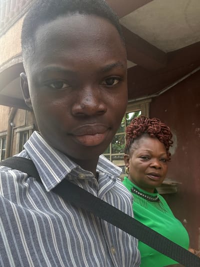 The image displays two individuals situated in Iriebe, Nigeria. In the foreground, a young, dark-skinned male with short, dark hair looks directly at the viewer. He wears a light blue and white vertically striped button-down shirt, with a black strap crossing his torso diagonally from his left shoulder. Behind him and to his right, an older, dark-skinned female is visible. She has reddish-brown twisted or braided hair, partially shaved at the sides, and wears gold hoop earrings. Her attire consists of a bright green top with a distinct black beaded neckline. She gazes generally towards the viewer. The background features a reddish-brown wall structure, a dark ceiling beam, and a window through which green foliage is visible.