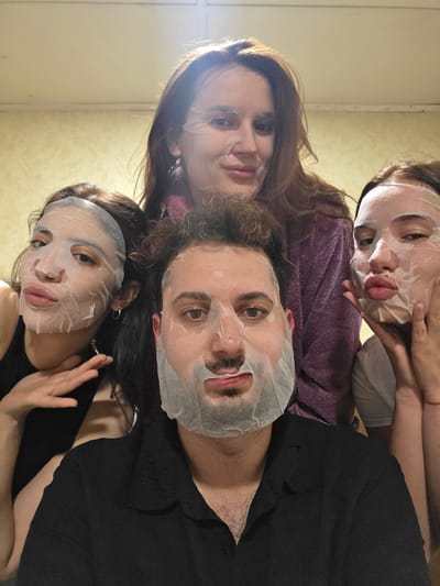 Four individuals, two men and two women, are posed for a selfie in what appears to be an indoor setting, possibly a residential room, in Aldomirovtsi, Bulgaria. All subjects are wearing white, translucent sheet masks on their faces. The woman in the center background, wearing a purple robe, is partially visible. A man in the foreground is wearing a black collared shirt. To his left, a woman in a black tank top is partially visible, gesturing with her right hand. To the right of the man, another woman is visible, wearing a white t-shirt and resting her chin on her hands. The background is a plain, textured wall.