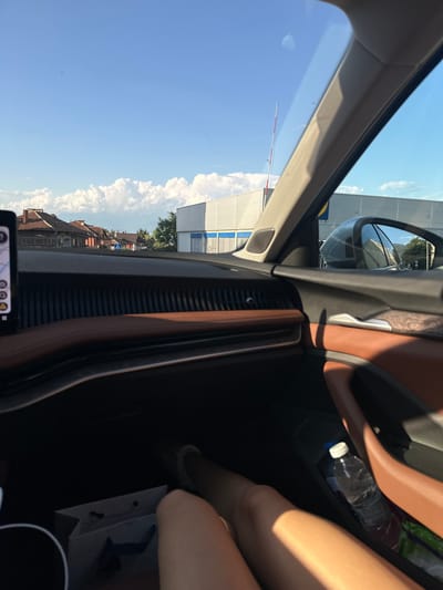 The image displays a detailed view from the interior of a vehicle, looking out through the front passenger window on a clear day in Petrich, Bulgaria. The sky is predominantly blue with scattered white cumulus clouds.

In the mid-ground, a row of traditional-style buildings with terracotta-tiled roofs is visible to the left. A blue and white pedestrian crossing sign is situated on the street in front of these structures. To the right, a large, modern commercial building with a grey paneled facade and blue lower sections is prominent, featuring a partially visible yellow and blue logo consistent with Lidl. A red and white striped barrier arm extends vertically near the center, positioned in front of this commercial establishment. Distant mountains are discernible on the horizon beneath the clouds.

The interior of the vehicle features a dashboard and door panel trimmed with dark brown leather and textured black plastic elements. A portion of a digital display screen is visible on the left side of the dashboard. A vehicle's side mirror is visible on the right, reflecting part of the commercial building and trees.

In the lower foreground, the bare legs and knees of a person, presumably the passenger, are visible. A plastic water bottle with a white cap is placed in a storage compartment within the passenger door, next to a green item. On the floor in the lower left, a white paper shopping bag with dark handles is present. The scene is illuminated by bright natural light.