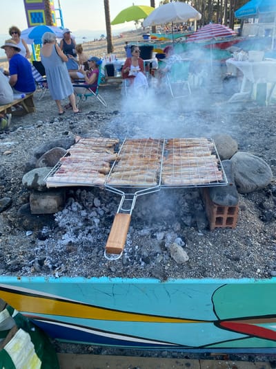 The image captures a lively scene on a beach in Marbella, Spain, possibly during a casual gathering. In the foreground, a barbecue grill with several rows of meat is actively cooking over hot coals, generating a visible plume of smoke. The grill is precariously balanced on rocks and a brick. Behind the grill, a group of people are enjoying themselves in a relaxed atmosphere. Some are seated in beach chairs, while others are standing and chatting. Umbrellas, some brightly colored, dot the sandy beach, providing shade. In the background, the calm sea and a distant coastline suggest a typical Mediterranean setting. The overall impression is one of a pleasant outdoor meal or celebration on a sunny day.
