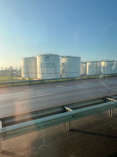 The image depicts a row of large white cylindrical storage tanks, likely for fuel or other industrial liquids, lined up behind a chain-link fence. The tanks are situated in an industrial area, with some piping and structures visible in the background. One of the tanks prominently displays the logo "Terminal" with the Estonian text "Eesti kütusepank" below it, indicating this is likely the Estonian Fuel Terminal. The foreground shows a wet asphalt road with white lane markings, suggesting it has recently rained or there is morning dew. A metal guardrail separates the road from the industrial area. The sky is clear and light blue, indicating it is likely early morning or late afternoon due to the soft lighting and long shadows. The overall scene is a snapshot of an industrial facility adjacent to a roadway, likely located in or near Maardu, Estonia, given the text on the tank.