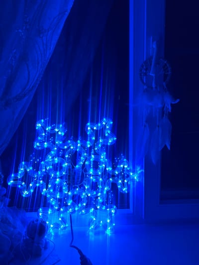 The image shows a blue LED snowflake decoration illuminated in a dark room. The snowflake is positioned on a windowsill, with a patterned blue curtain to the left and a window frame to the right. A dreamcatcher with white feathers hangs from the window frame. The overall scene is bathed in the bright blue light of the decoration, with some ambient blue light reflecting off surfaces. The context provided about Krasnoe-na-Volge, Russia, is not visually supported by the image itself.