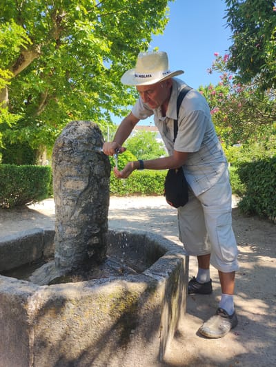 A man wearing a straw hat and a light-colored, short-sleeved, plaid shirt is standing next to a stone fountain in a park. He is leaning over the fountain, with his hands under the running water from the tap. He is wearing light-colored shorts, white socks, and dark sneakers. A dark bag is slung over his shoulder. The fountain is made of rough, weathered stone and has a stone basin. The scene is outdoors, with lush green trees and bushes surrounding the fountain. Pink flowers are visible in the background. The sky is bright blue and clear, indicating a sunny day. The sunlight casts shadows on the ground and on the fountain. The man appears to be refreshing himself with the water. The hat has text on it that reads "TERRA MISLATA". The setting appears to be a park or garden in a warm climate.