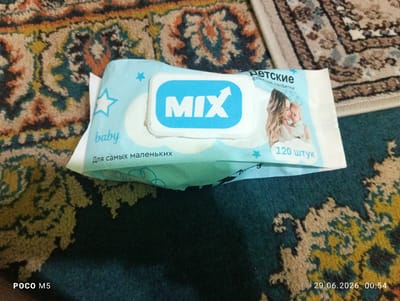 A pack of "MIX" baby wet wipes is shown on a patterned rug. The pack is light blue with white and turquoise accents and features a logo with the word "MIX" in large, bold letters. Text on the pack includes "ДЕТСКИЕ" (Children's) and "Влажные салфетки" (Wet wipes). Below the logo, the text "baby" is visible in a turquoise font, and further down, "Для самых маленьких" (For the smallest ones) is printed. A portion of an illustration of a woman holding a baby is also present. The bottom right corner of the image displays the date "29.06.2026 00:54" and "POCO M5". The background is a rug with a complex, multicolored pattern. There are no discernible elements that place this image specifically within the city of Hisor, Tajikistan.