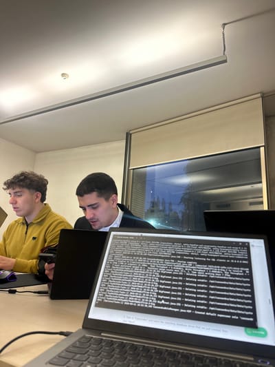 The scene depicts an interior workspace within the city of Guimarães, Portugal. Two young adult males are seated at light-colored desks, engaged with individual laptop computers.
The individual positioned on the left has short, curly dark brown hair and is wearing a mustard yellow long-sleeve polo shirt with a small dark emblem on the left chest. His gaze is directed downwards, and his right hand rests on a black computer mouse situated on a dark mousepad that exhibits a subtle purple LED glow.
The individual in the center has short, dark hair and is wearing a dark jacket over a white collared shirt. He is looking down at a black laptop screen directly in front of him.
In the foreground, a silver-colored laptop is visible, its screen displaying an active terminal interface with technical text. The content includes references to "Cassandra" and "Java," suggesting programming or system administration activities. A black cable is connected to the left side of this laptop.
The environment includes a white ceiling with recessed linear lighting fixtures and a white circular smoke detector. A coiled wire descends from the ceiling near the windows. Large windows line the background, partially covered by beige roller blinds. Reflections of the interior lighting are visible on the glass, behind which a dark, indistinct outdoor scene featuring trees and distant lights is discernible, indicating evening or nighttime conditions. The walls are light-colored.