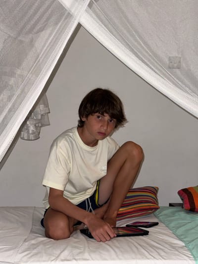 A boy sits cross-legged on a bed with his head propped up. He wears a light-yellow t-shirt and dark shorts. He is looking at the camera with a slight, quizzical expression. He holds two closed, black tablets in his hands, near his feet. A striped pillow sits behind him. A mosquito net hangs around the bed. The scene is dimly lit.