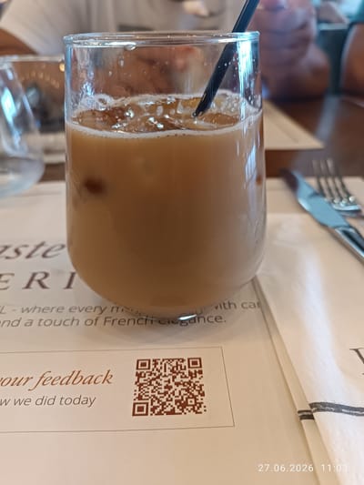 A close-up shot shows an iced coffee drink in a clear glass, filled with ice cubes and a light brown liquid. A black straw is visible in the drink. The glass sits on a white paper, likely a placemat or menu, which has some text printed on it. To the right of the glass, a white napkin is folded, and a silver knife and fork are placed on top of it. In the background, out of focus, are hints of people and a blurry glass. The scene suggests a dining or cafe setting. A timestamp "27.06.2026 11:03" is visible in the bottom right corner.
The text on the placemat is partially visible and reads: "ste", "ERI", "L - where every m", "and a touch of French elegance.", "our feedback", and "w we did today". A QR code is also printed on the placemat. The overall mood appears casual and relaxed, with the bright lighting suggesting it's daytime. The location context provided is Strovolos, Cyprus.
