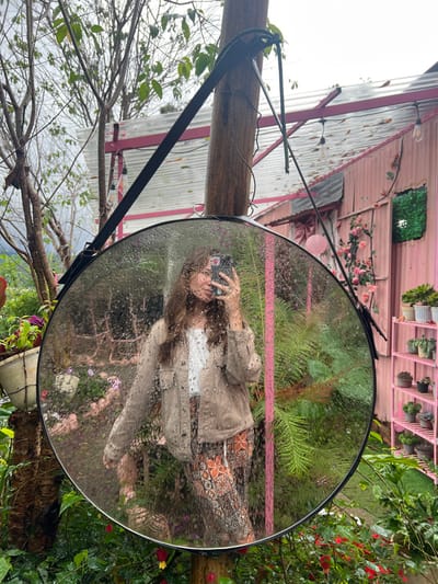 The image presents an outdoor scene located in Đưng K'Si, Vietnam. A large, circular mirror, approximately 0.8 meters in diameter, with a black frame and covered in water droplets, is centrally positioned. It hangs from a stout wooden pole via a black strap. The mirror reflects a female subject, observed from the waist up. She possesses long, wavy brown hair, wears a light brown or beige button-up jacket over a white undershirt, and patterned pants featuring orange and dark motifs. The subject holds a black smartphone to her face, capturing her reflection, with a metallic ring visible on her right hand's ring finger.

The surrounding environment consists of lush, dense greenery. To the left, a tree with thin, leafless branches extends upwards against an overcast, grey sky. A white hanging pot with green foliage is attached to a smaller tree trunk. Behind the mirror, various green plants, including ferns, are visible. To the right and in the background, a distinctive structure constructed of pink corrugated metal is present. This building features a pitched roof with exposed pink support beams, and is decorated with string lights containing Edison-style bulbs. A large pink bow and artificial pink roses adorn its facade. A square opening in the wall is filled with green artificial foliage. Adjacent to the building on the right, a pink multi-tiered shelving unit displays an array of potted plants, including succulents and small bonsai trees. Red and green flowering plants are observed in the immediate foreground, contributing to the vibrant garden setting.
